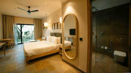A room with a king size bed with white bedding, a large vertical oval mirror on the wall, and a window overlooking the greens at Jivana Wellness Retreat, Bhopal.