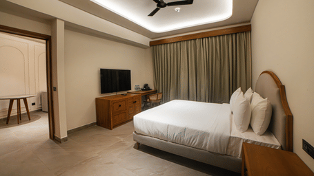 A suite with a king size bed, a television on a wooden stand, a ceiling fan, and long curtains covering a window at Jivana Wellness Retreat, Bhopal.