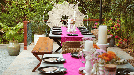 An outdoor dining setup with a long table, tableware, candles, floral arrangements, and a decorative hanging chair surrounded by lush greenery.