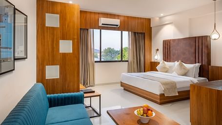 A well-lit hotel suite features a comfortable bed, a sofa, a wooden coffee table with fruit, and a window with curtains - Lords Eco Inn Ambaji.