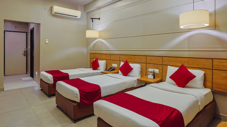 Three single beds in a hotel room with white bedding, red accents, a wooden headboard, air conditioning, and wall-mounted lamps.