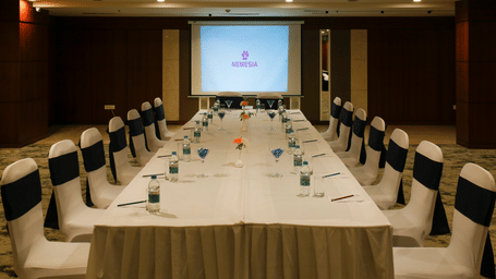 Long boardroom table set up for a corporate meeting, with chairs featuring navy sashes facing a large projector screen at the end of the room.