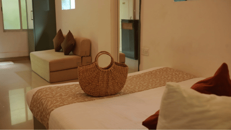 A bedroom at Oceanature Resort, Alibaug featuring a queen size bed with a woven bag, a sofa in the background, and mirrors.