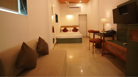 A bedroom at Oceanature Resort, Alibaug with a sofa, awall-mounted television, a wooden desk with chairs, and a queen bed in the background.