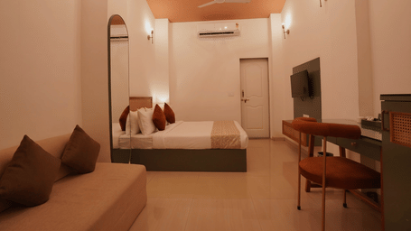 An interior view of a bedroom featuring a queen size bed with pillows, a low sofa in the foreground, and a wooden desk.