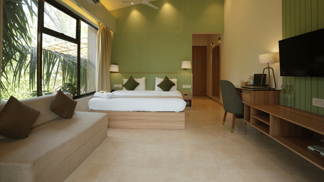A bedroom at Oceanature Resort, Alibaug with a king size bed against a green wall, a sofa, a long desk, work desk, chair, a wall-mounted television, and large windows.