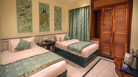 Diagonal view of the spacious room with twin beds placed side by side, a wooden bedside table in the middle, three photo frames above the headboard, and wooden storage | Panna Vilas Udaipur