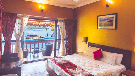 Room with a queen size bed, ceiling fan, framed picture, and view of the river through the window at Paradise Lagoon Resort, Udupi.