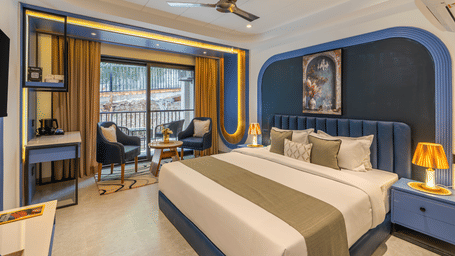 A bedroom featuring a king size bed with a blue headboard, side table with a lamp, work table, a ceiling fan, and a seating area near a sliding glass door at Perfectstayz Luxe Mall Road.