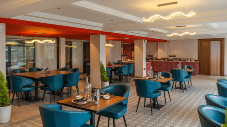 An indoor restaurant with multiple teal armchairs and wooden dining tables under modern light fixtures at Perfectstayz Luxe Mall Road.