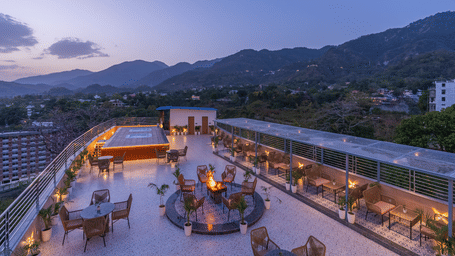 An aerial view shows an open-air rooftop with multiple seating areas and a glowing fire pit at dusk at Perfectstayz Premium Aurevva.