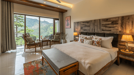 A room features a white bed, a grey footbench, and a sitting area by a large window with grey curtains at Perfectstayz Premium Seven Oaks, Mussoorie.