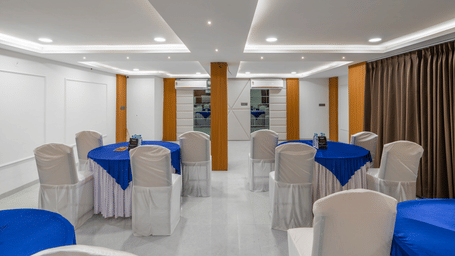 A well-lit hall at Perfectstayz Premium Shree Narayana, Ujjain featuring white chairs, blue table settings, and modern recessed ceiling lights.