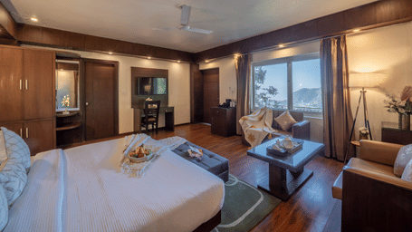 A bedroom at Perfectstayz Premium The Pride, Mussoorie with a king size bed, seating area with 2 sofas, window view, wardrobe with a vanity and side tables.