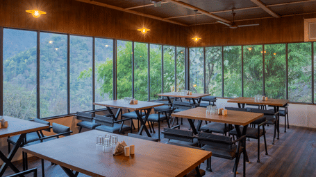 A restaurant area at Perfect Stayz The Jungle Resort has tables, chairs, and glass walls overlooking nature.