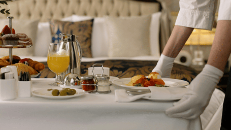 Two hands placing a bowl of fruits on a table filled with eateries near a bed.