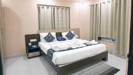 Bedroom of an Executive Suite at Pipul Hotel And Resort, Puri, featuring a plush bed with decorative towel art, patterned cushions, a nightstand, and draped windows.