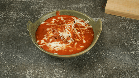 Creamy tomato-based curry topped with grated cheese, served in a stoneware bowl on a textured grey surface beside a wooden board.