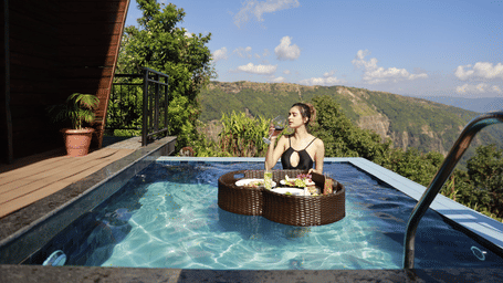 Woman in black swimsuit at Polo Resort Cherrapunji private plunge pool with floating breakfast tray, sipping a drink overlooking cliffs and green hills.