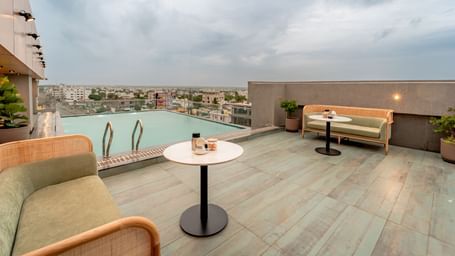 The rooftop swimming pool with clear water, surrounded by a tiled deck and some modern outdoor seating, offering a panoramic view of a city under a cloudy sky | Pravasa Hotel Vijayapura