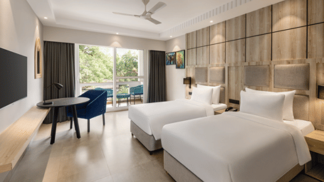 Bright hotel room with twin beds, work desk and a private balcony overlooking greenery.