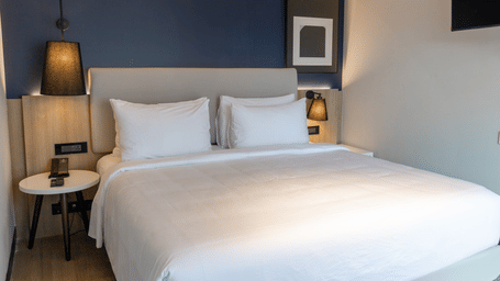 A neatly dressed king bedroom in the Sands Point Hotel Deluxe Suite featuring a large bed with a light grey upholstered headboard, crisp white pillows and linen, built-in wooden bedside panels with black swing arm reading lamps, a round white bedside table