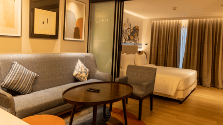 A wide-angle view of the Executive Suite at the Sands Point Hotel, Pondicherry, showing the layout with the living area featuring a grey sofa with cushions, a round dark coffee table, and framed artworks on the wall.