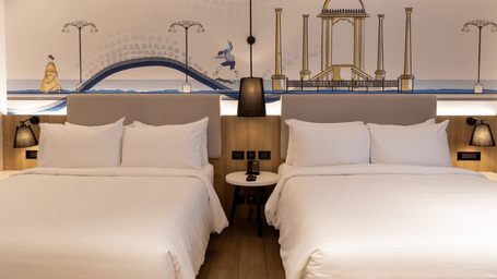 A close-up view of the 2 beds in the Family Room at Sands Point Hotel, Pondicherry, each in white linen with pillows and light grey headboards with warm integrated lighting, and shared bed lamps.