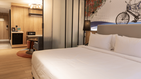 A wide view of the Sands Point Hotel Luxury Suite bedroom showing a king bed with white linen and a grey upholstered headboard, a black pendant lamp, a frosted glass sliding partition partially open to reveal the living and amenities area beyond with a woo