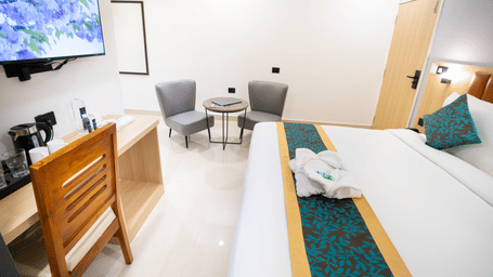 A view of the room with a large TV, a small table with chairs to the side, wooden interiors, and a spacious bed at Sunrise Business Hotel, Hyderabad.