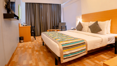A bedroom at TGI Ameya Grand, Panjim, Goa, featuring a bed with a colourful runner, a solid headboard, a flat-screen TV, and a seating area. 