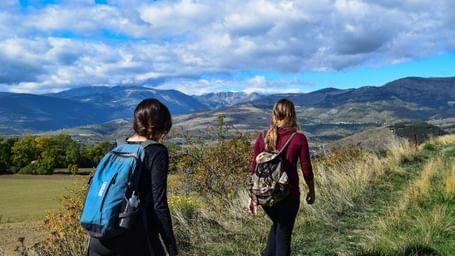 Two women with backpacks hiking along a grassy trail overlooking scenic mountains, valleys, and a partly cloudy blue sky.
