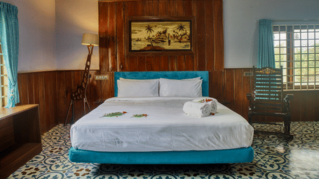 A hotel bedroom featuring a large bed with white linens and a blue bed frame, a wooden headboard, and a window with light filtering in.