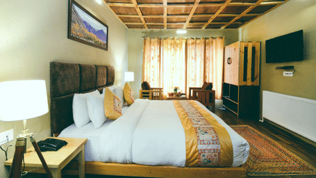 Standard Cottage featuring a king size bed with white linens and a decorative throw, wooden furniture, and a ceiling with a geometric pattern at White Sands Resort, Ladakh.