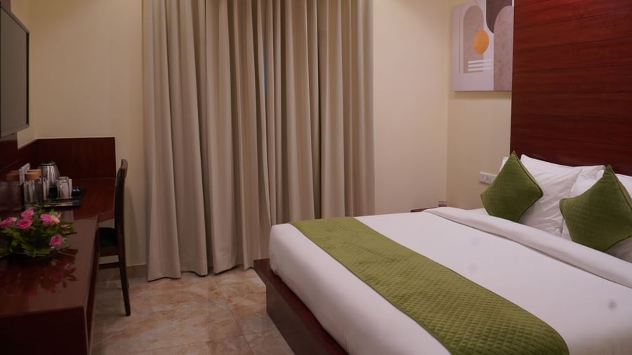 A wide shot of a hotel room with a TV on a wooden wall panel, a desk, a chair, and a bed with a white comforter and a green accent runner.