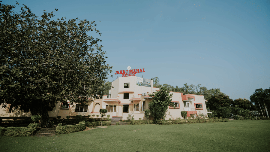 A facade view across a large green lawn looking towards the building exterior of TGI Jahaj Mahal, Mandu, partially framed by trees.