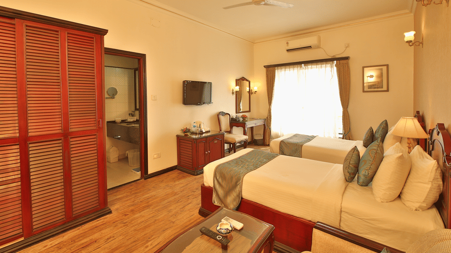 Hotel guestroom featuring double beds, a wooden wardrobe, television, and window curtains at The Lindsay, Kolkata.