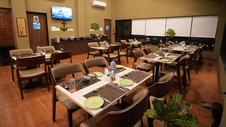 A restaurant with neatly arranged tables and chairs, offering a warm and inviting, dimly lit atmosphere. It features a television on the wall, air-conditioning units, and potted plants for added charm - Gaurav Lords Resort Shrivardhan.