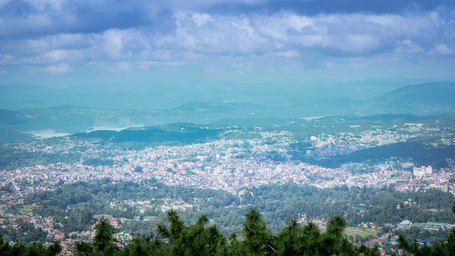 Aerial view of a dense hillside town with clustered buildings, forested hills, a distant lake, and a partly cloudy blue sky.