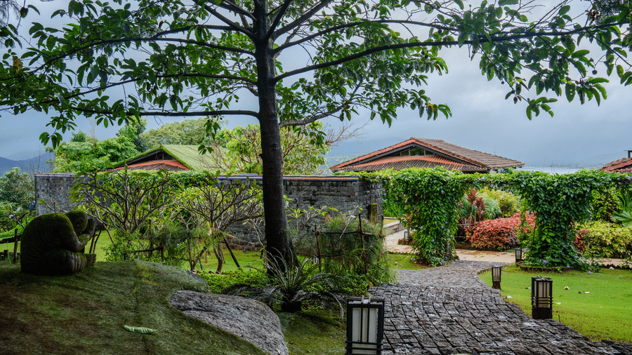 A cobblestone pathway winding through a lush green resort garden with a large tree and stone walls under a cloudy sky.