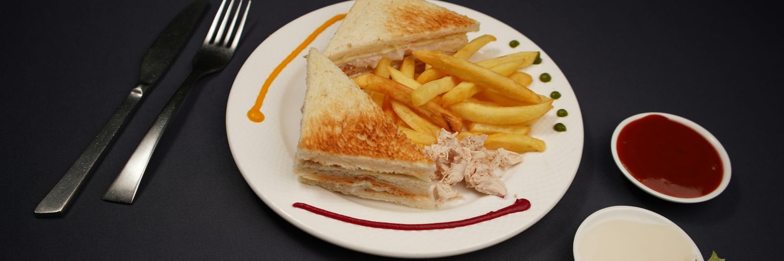 A plate of sandwich, is served with dipping sauces and a lime wedge on a dark surface.