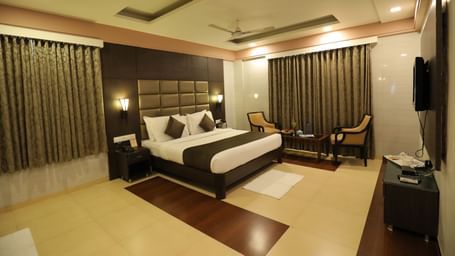 A front angle view of a bedroom at Krushnai Resort with a king size bed, a dark headboard, and brown drapes.