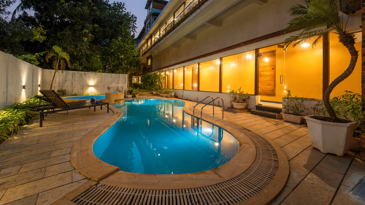 Night view of Rosetum Anjuna's outdoor pool with glowing hotel facade, multi-storey balconies, sun loungers, and tropical trees in the background.