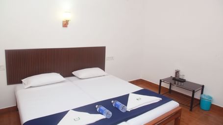 A bedroom with white walls, a double bed with blue runner, towels and water bottles neatly placed  on them at Hotel Srinivas, Kochi.