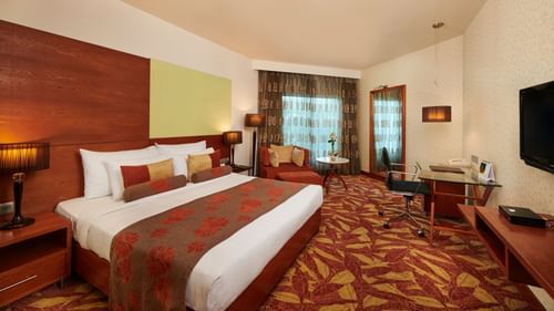 Room at Pride Premier Nagpur featuring a plush bed with pillows and cushions placed in front of a TV and a sitting space next to the window