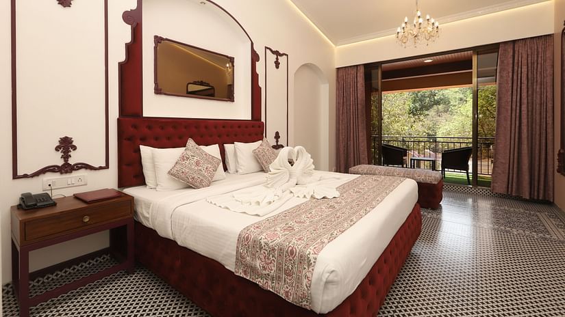Moghul Room at Zaras Resort with elegant décor, red accents, swan towel art on bed, balcony view of greenery, and ambient lighting.