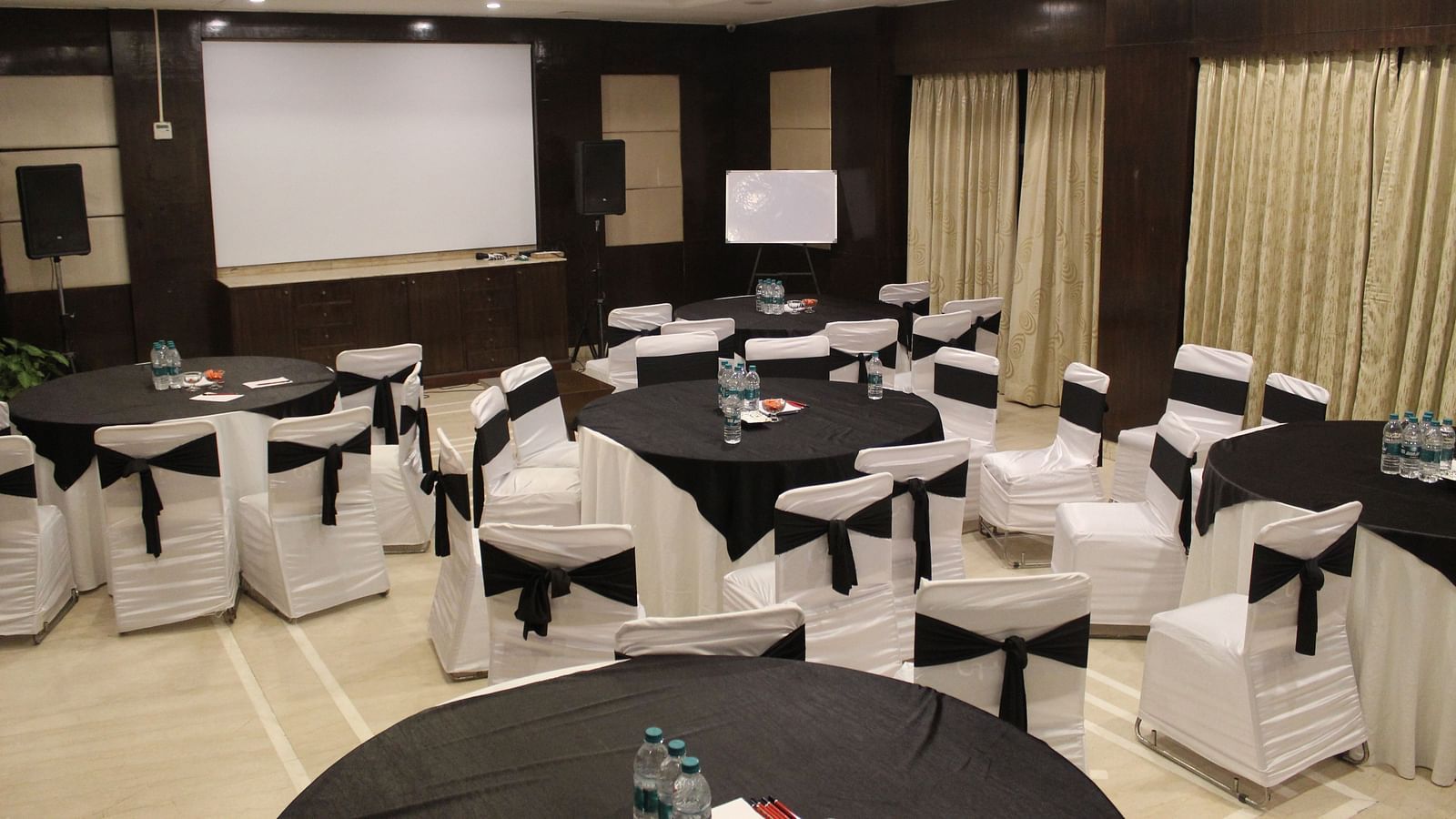An image of a banquet hall at Inde Hotel Signature Tower, Gurgaon featuring a few round tables, each surrounded by chairs
