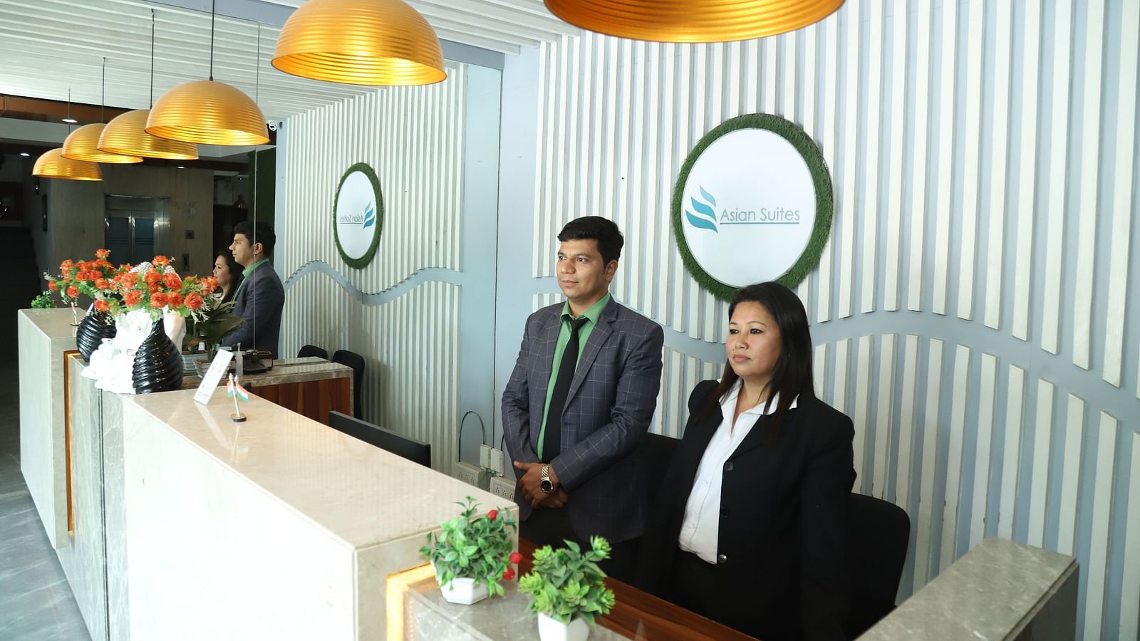 Two individuals stand behind a reception desk in a well-lit area with decorative lighting - Asian Suites IFFCO Metro, Gurgaon