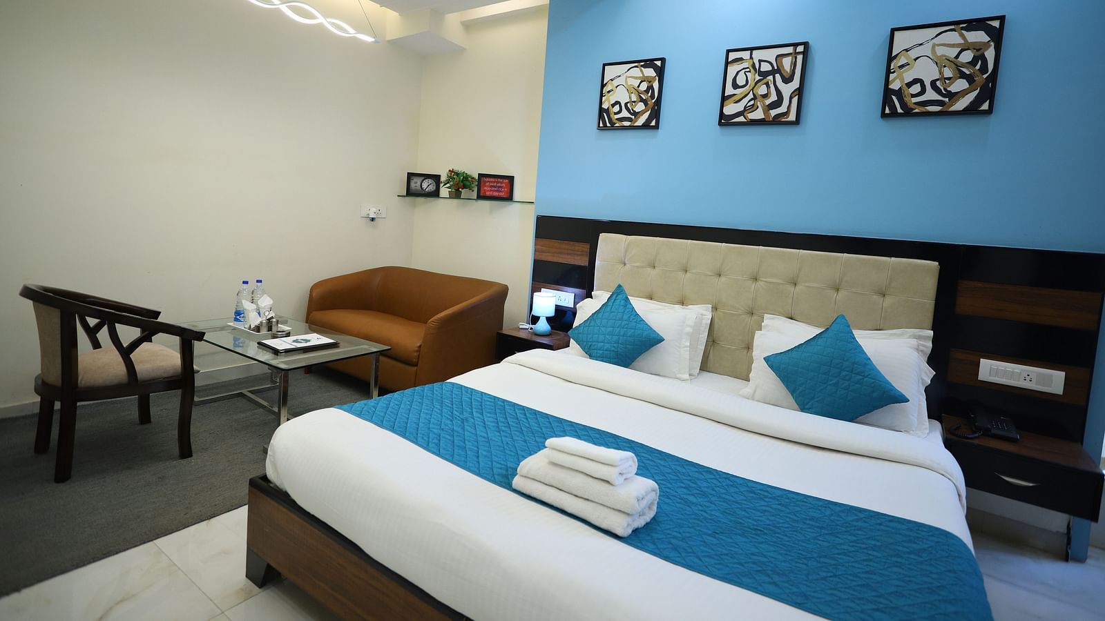 A comfortable room features a double bed with blue and white bedding, a seating area, and framed pictures on the wall - Asian Suites Cyber Park, Gurgaon