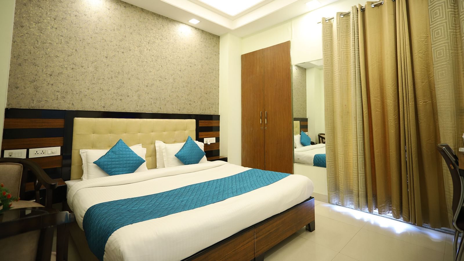 A well-lit room includes a double bed with blue accents, wooden furniture, and a mirror on the wall - Asian Suites Cyber Park, Gurgaon